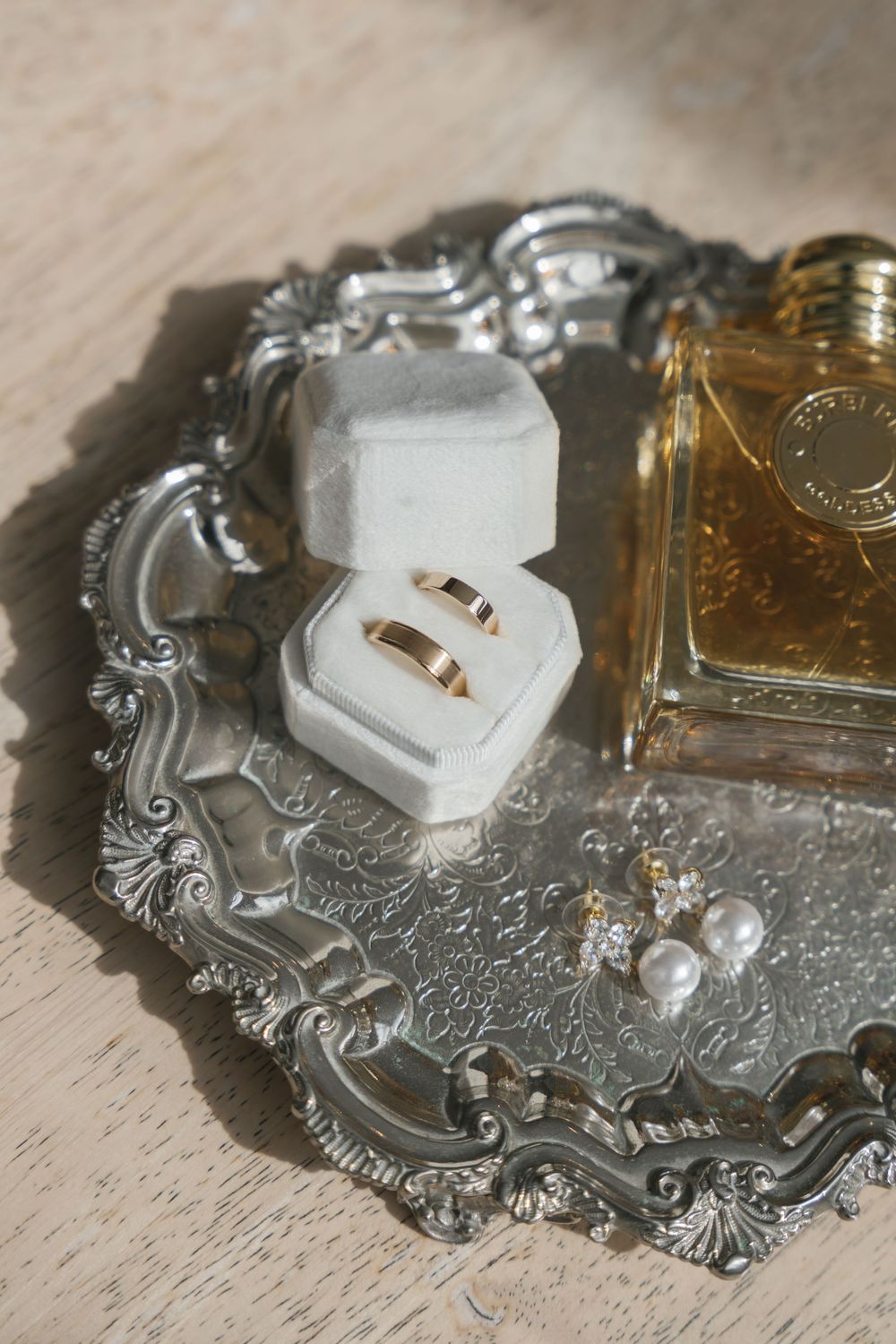 Two gold wedding bands displayed in a white hexagonal ring box on an ornate silver tray with perfume and earrings.