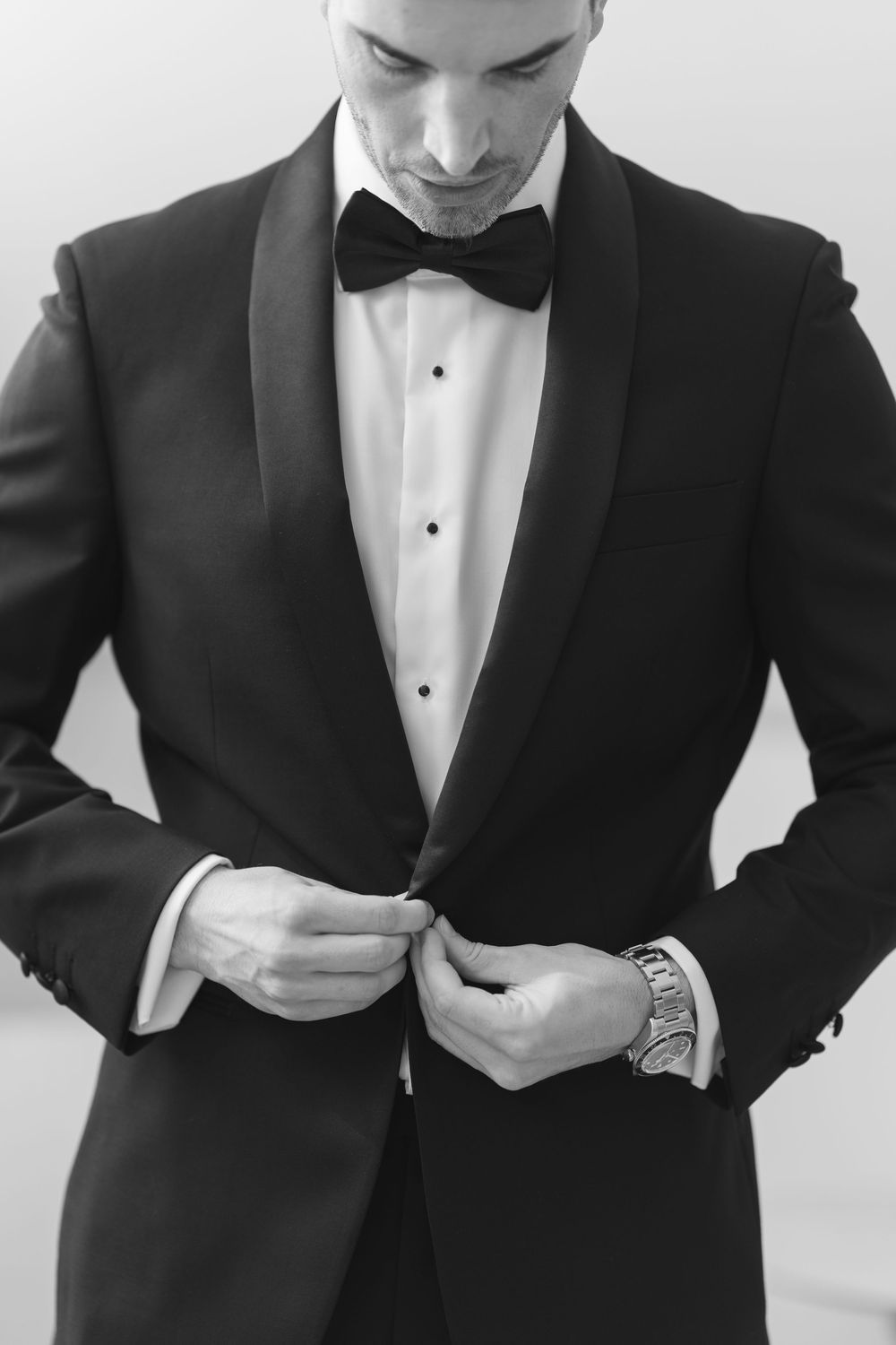 Man in black tuxedo with bow tie buttoning jacket, black and white formal portrait photo.