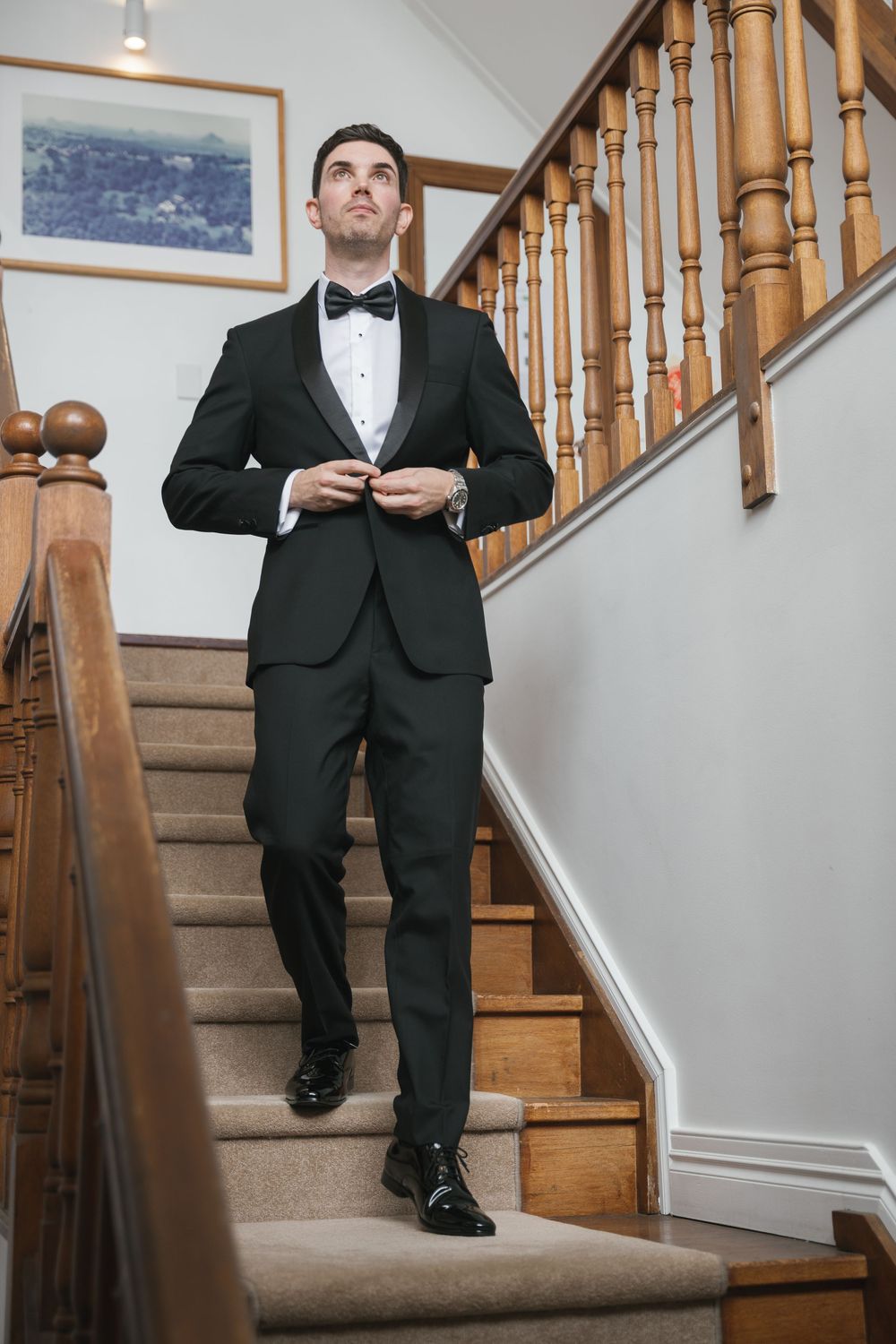 Elegant groom in black tuxedo and bow tie buttoning jacket while descending wooden staircase indoors.