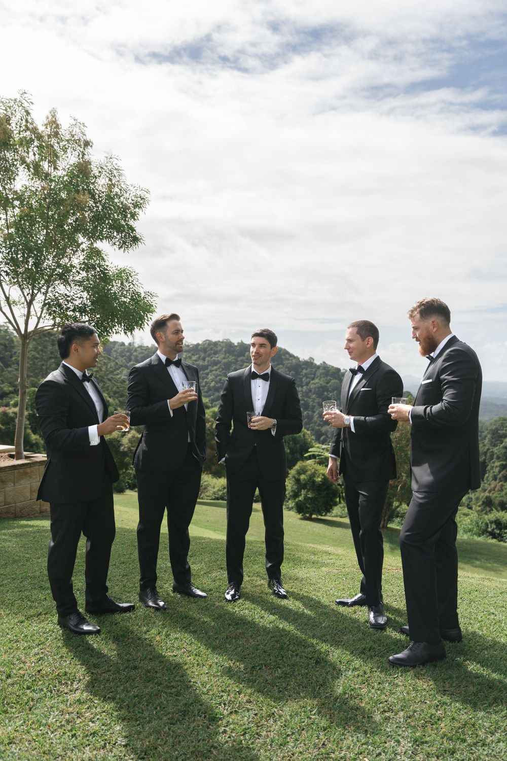 Five groomsmen in black tuxedos holding champagne glasses on a lush green lawn with scenic mountain views.