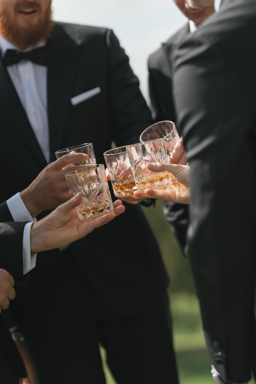 Men in black tuxedos clinking crystal whiskey glasses outdoors at a formal celebration or wedding toast.
