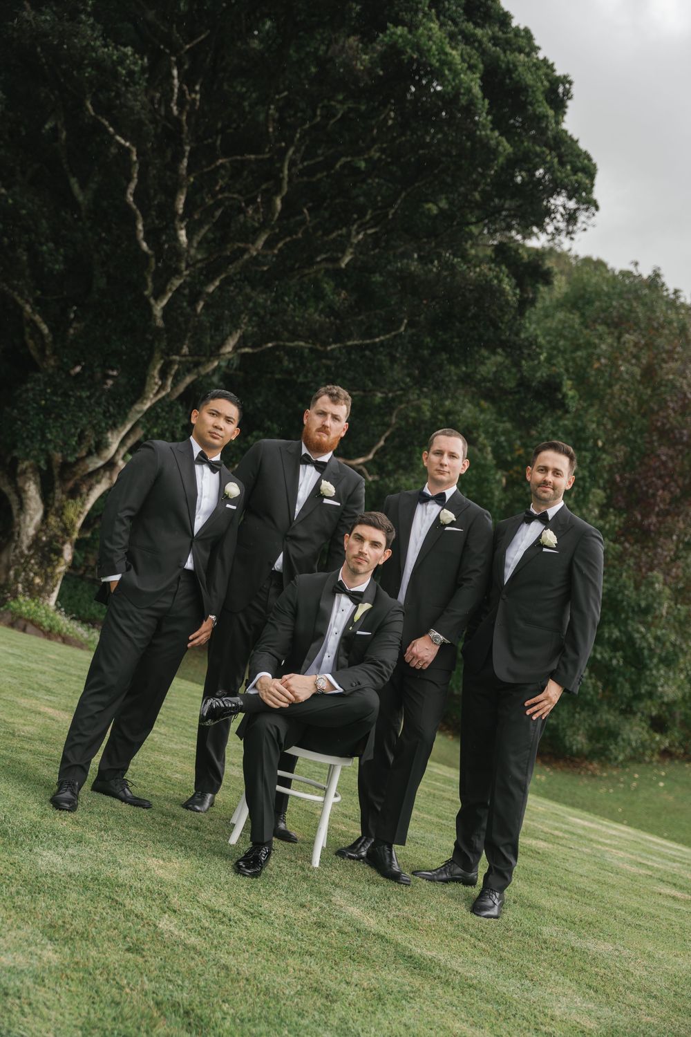 Five groomsmen in matching black tuxedos with bow ties pose outdoors on green grass for a wedding photo.