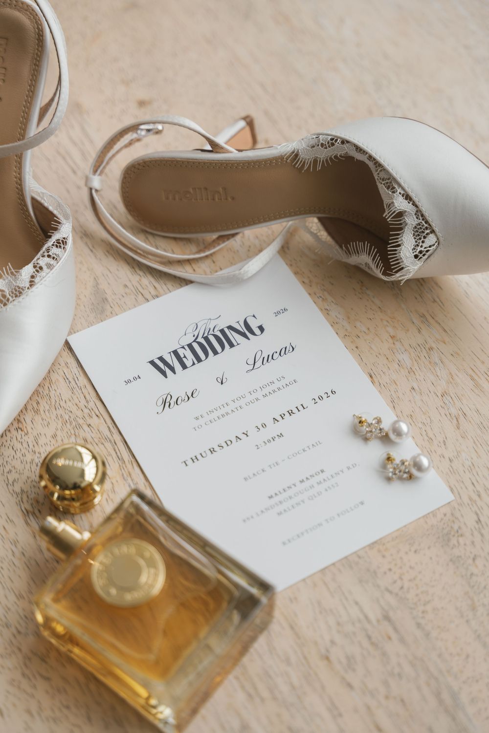 Wedding flat lay with white heels, floral invitation for Rose and Lucas, pearl earrings, and gold perfume bottle on wood surface.