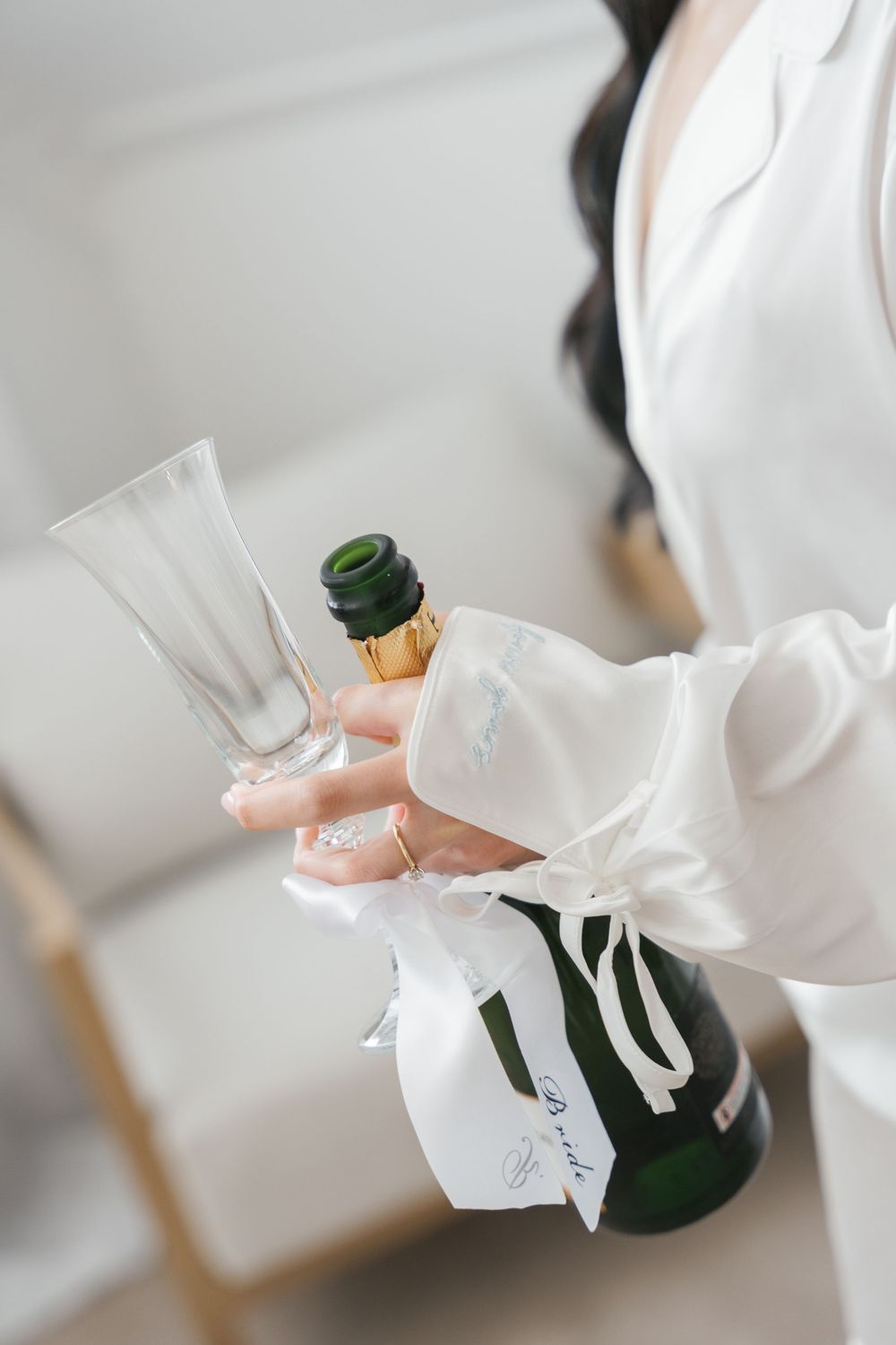 Bride in white robe holding champagne flute and decorated bottle with ribbon and tag.