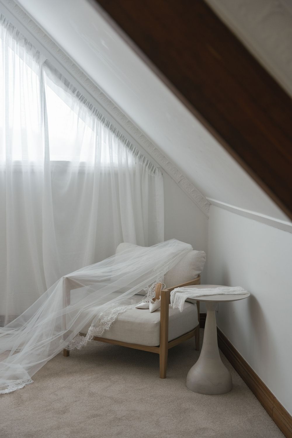 Elegant white wedding veil draped over a chaise lounge in a bright attic bridal suite with sheer curtains.