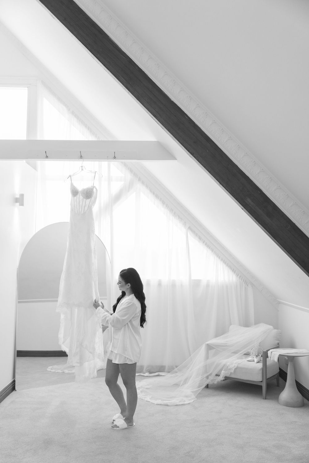 Black and white photo of bride in white robe admiring her wedding dress hanging in a sunlit attic room.