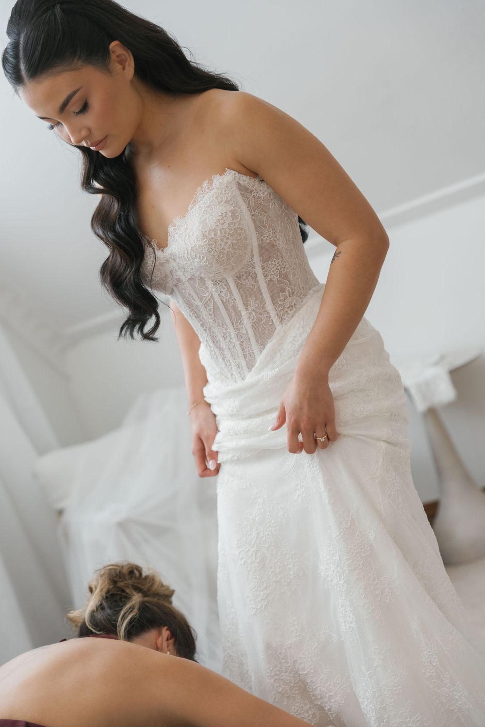 Bride in strapless lace wedding gown being helped with her dress by a bridesmaid in a bright bridal suite.
