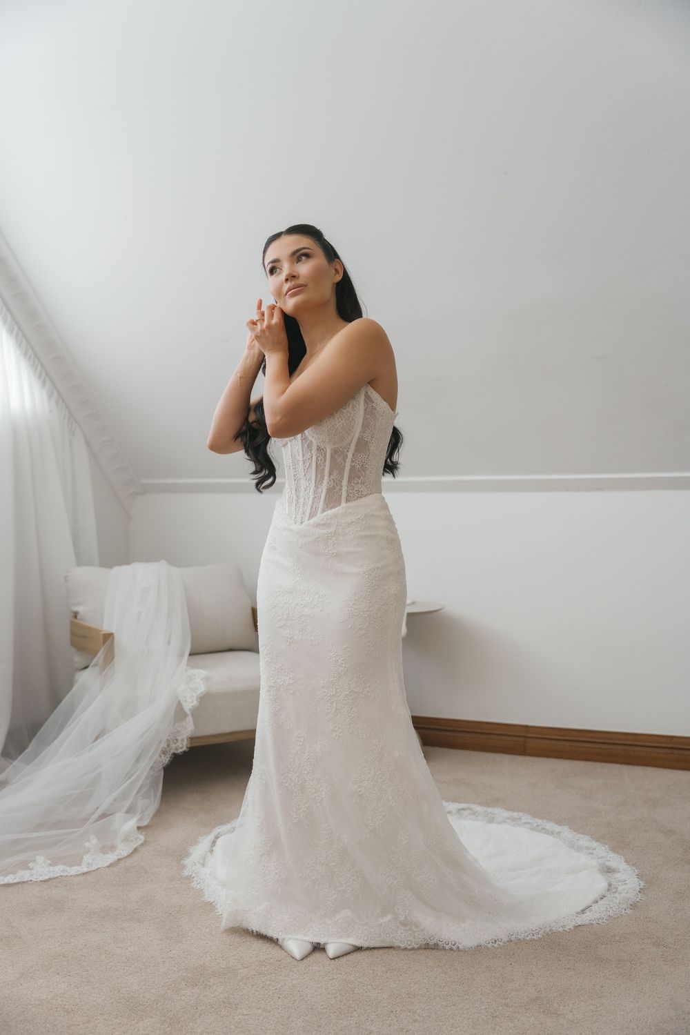 Bride in a fitted lace corset wedding gown with train poses elegantly in a bright white bridal suite.