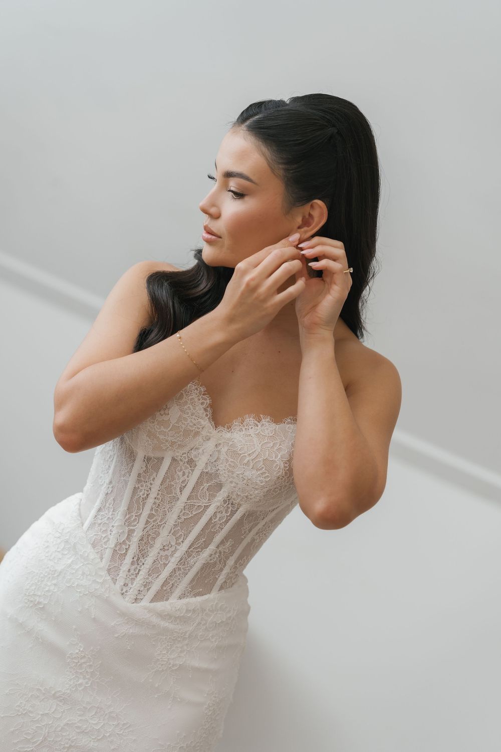 Bride in lace corset wedding dress adjusting earring against white wall.