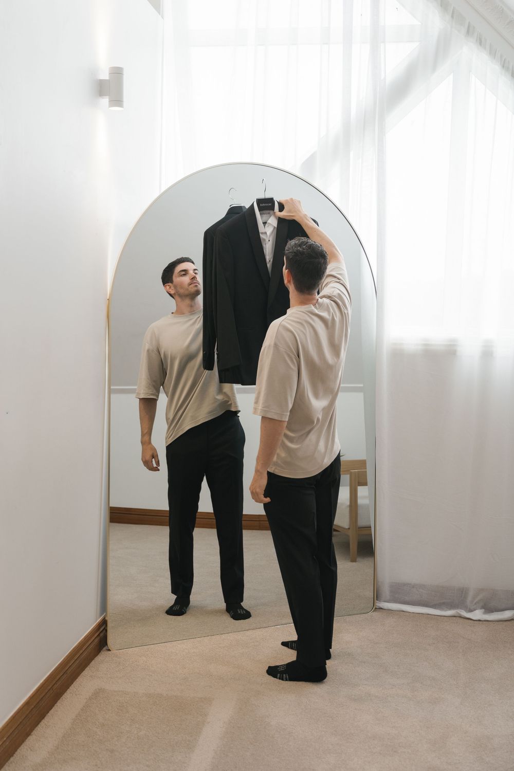 A groom holds a black tuxedo jacket up to a large arch mirror while getting ready for his wedding day.