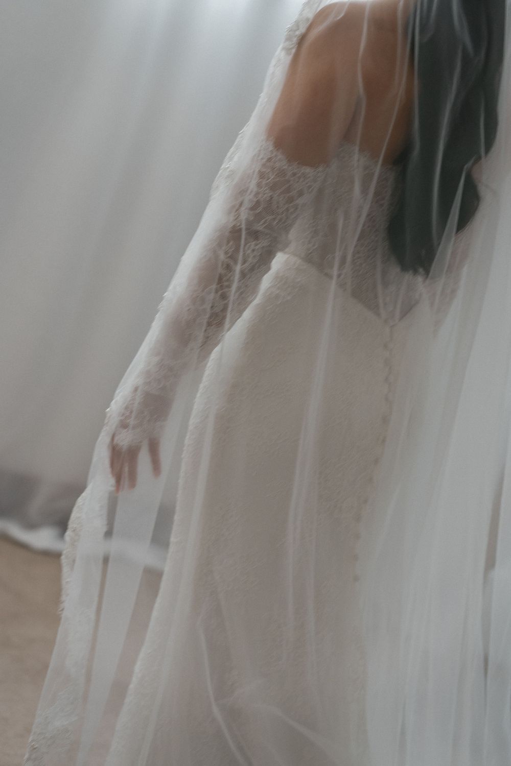 Elegant bride in long-sleeve lace wedding dress draped with a flowing cathedral veil in soft light.