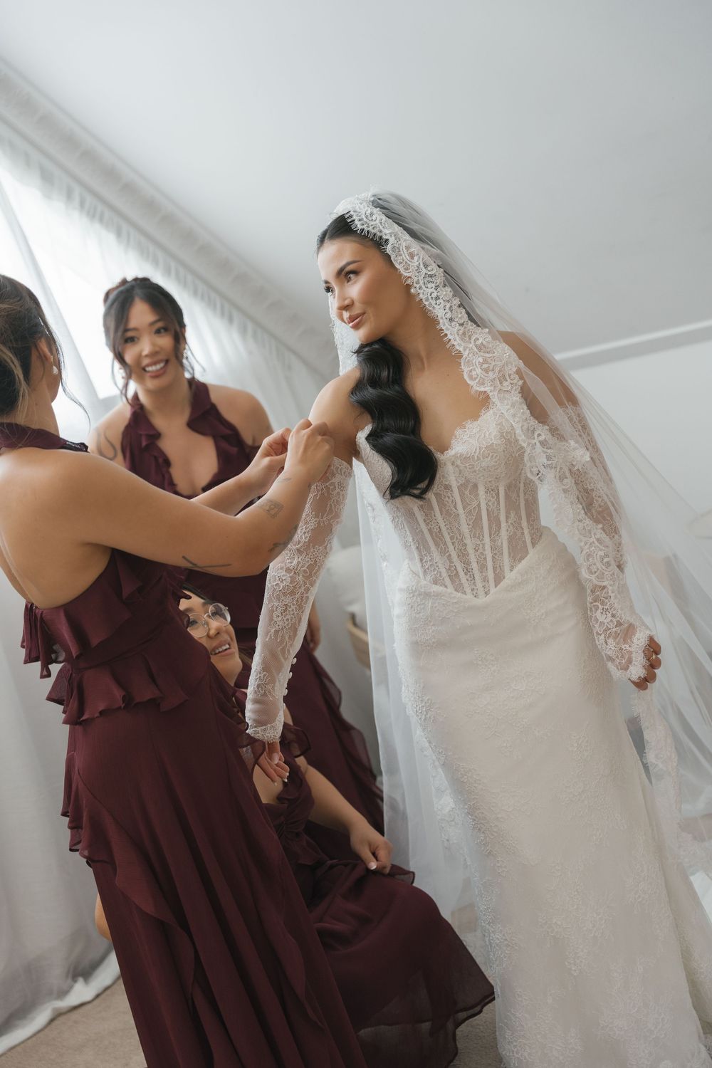 Bridesmaids in burgundy dresses help bride in lace wedding gown and veil prepare for her wedding day.