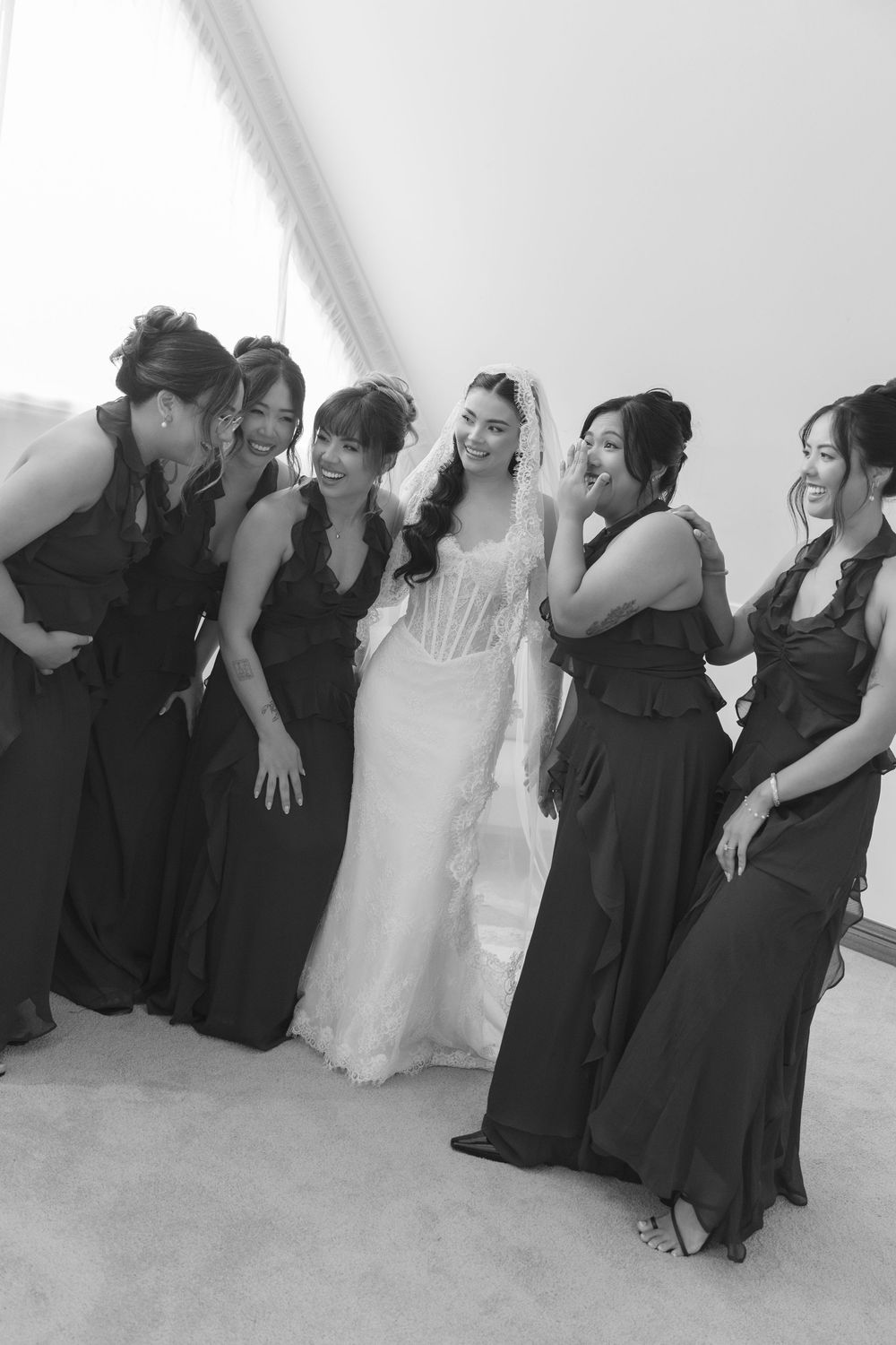 Bride in lace gown laughing with bridesmaids in black dresses, black and white photo.