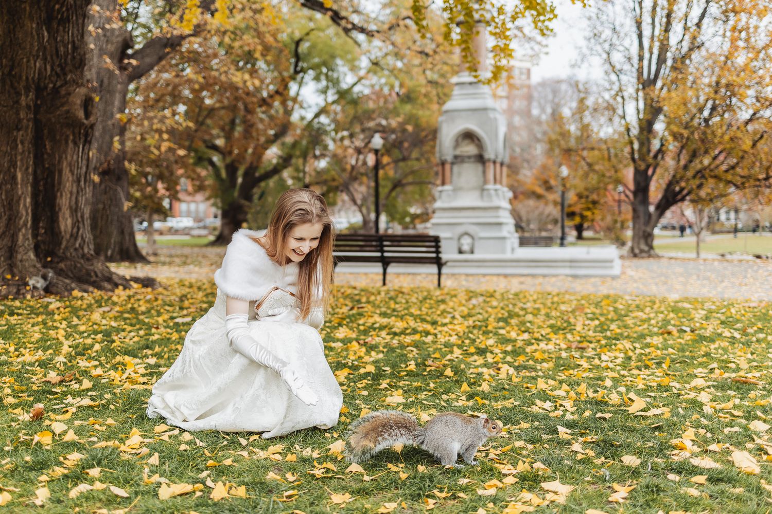 Girl in white dress crouching to feed a squirrel in an autumn park with yellow leaves and a monument.