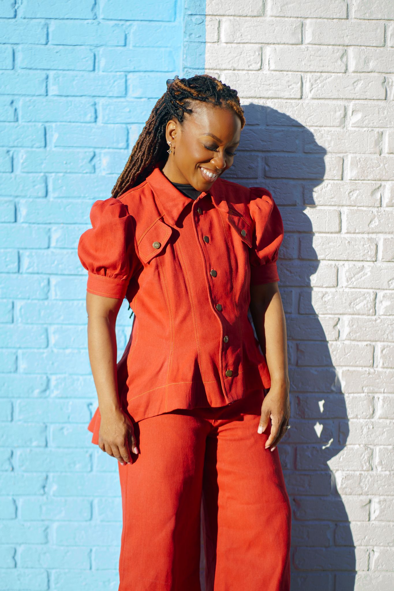 Smiling woman in red puff-sleeve top and wide-leg pants posing against a blue and white brick wall.