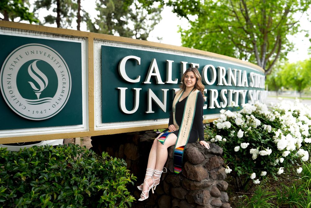 Graduate poses in front of California State University sign surrounded by white flowers and greenery.