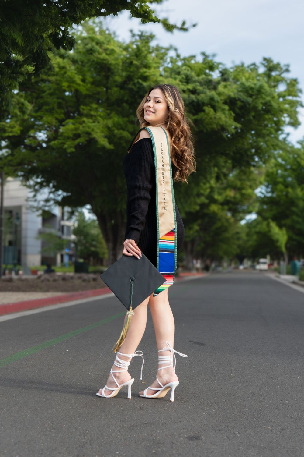 Young woman in graduation attire with colorful stole holds cap while posing on street with trees.
