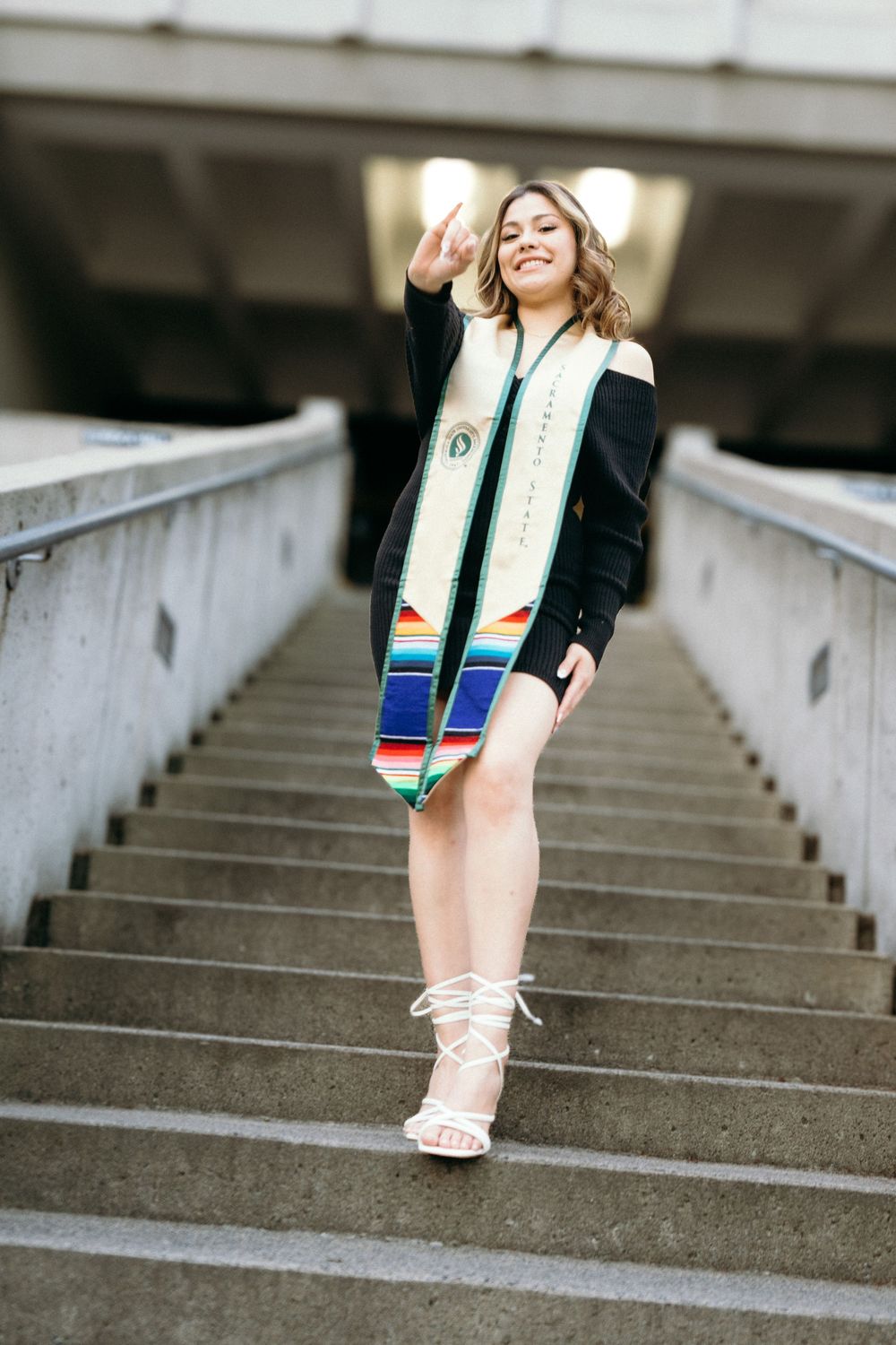 Smiling graduate in black outfit with colorful stole poses on outdoor staircase in white heels.