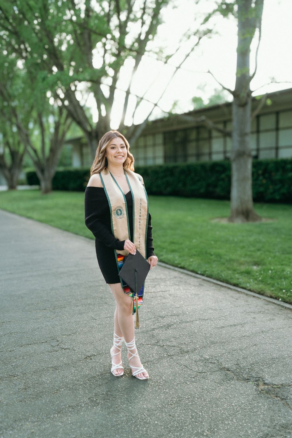 Young woman in graduation attire holding cap, wearing black dress and white heels, posing outdoors on a tree-lined path.