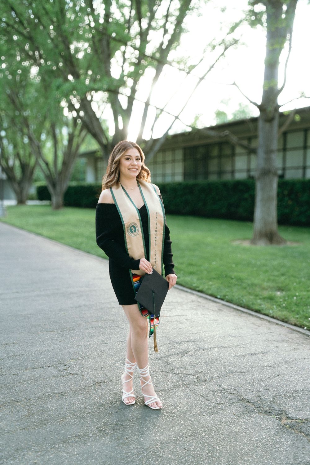 Young woman in graduation attire holding cap and stole, smiling outdoors on a tree-lined path.