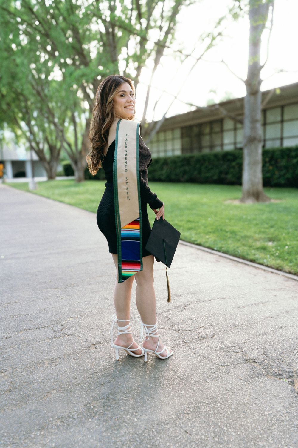 Smiling graduate in black dress with colorful sash holds cap outdoors on campus pathway.
