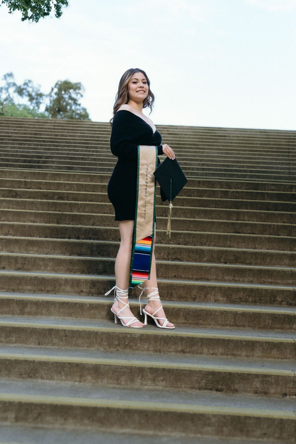 Young woman in black dress holding graduation cap and colorful stole on outdoor stone steps.
