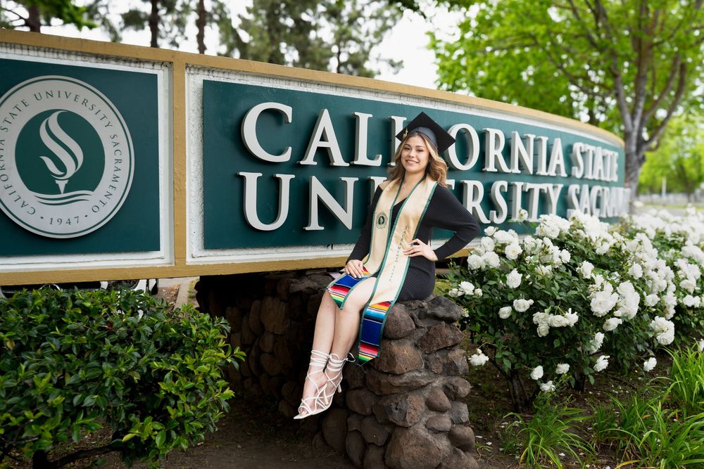 Graduate poses in cap and gown by California State University Sacramento sign surrounded by white flowers.