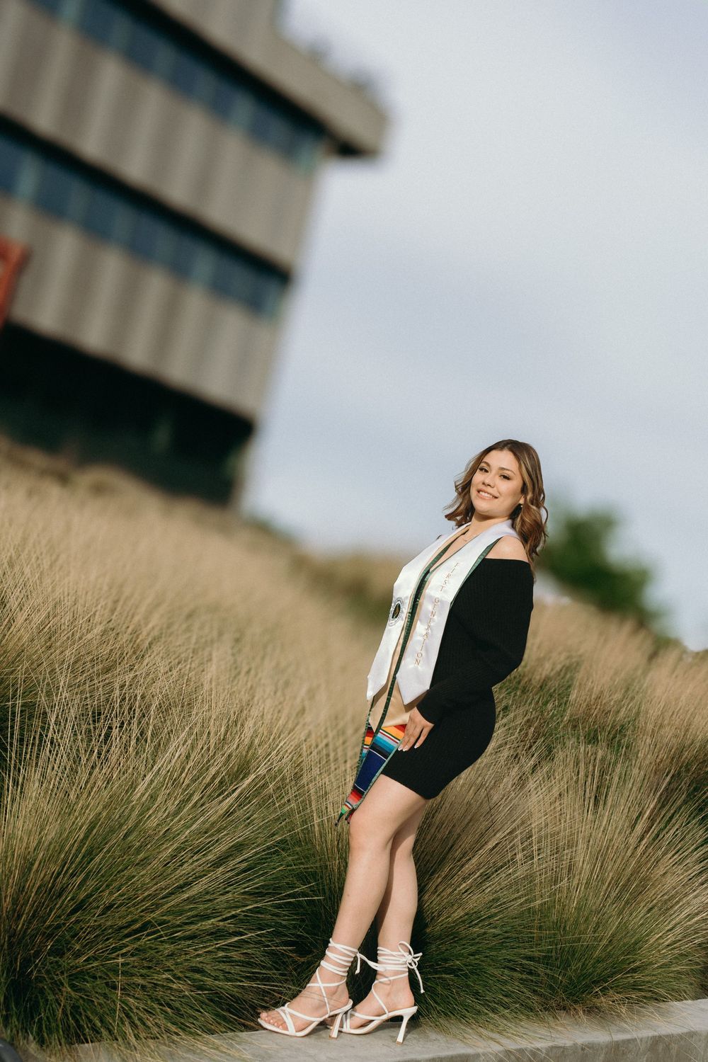 Young woman in black dress and white graduation stole smiles outdoors near tall grasses and modern building.