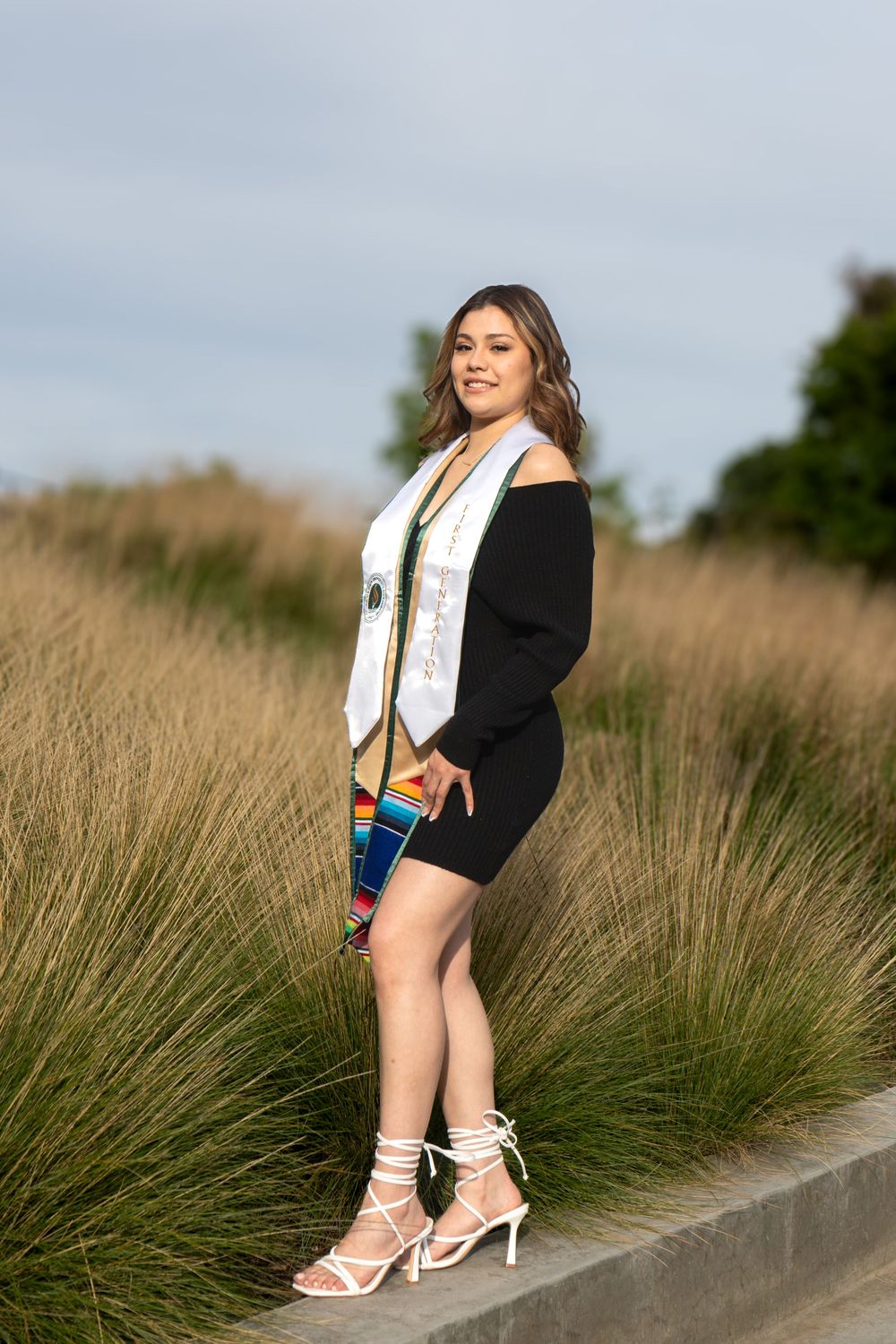 Young woman in black dress and colorful graduation stole poses outdoors among tall grasses in white strappy heels.