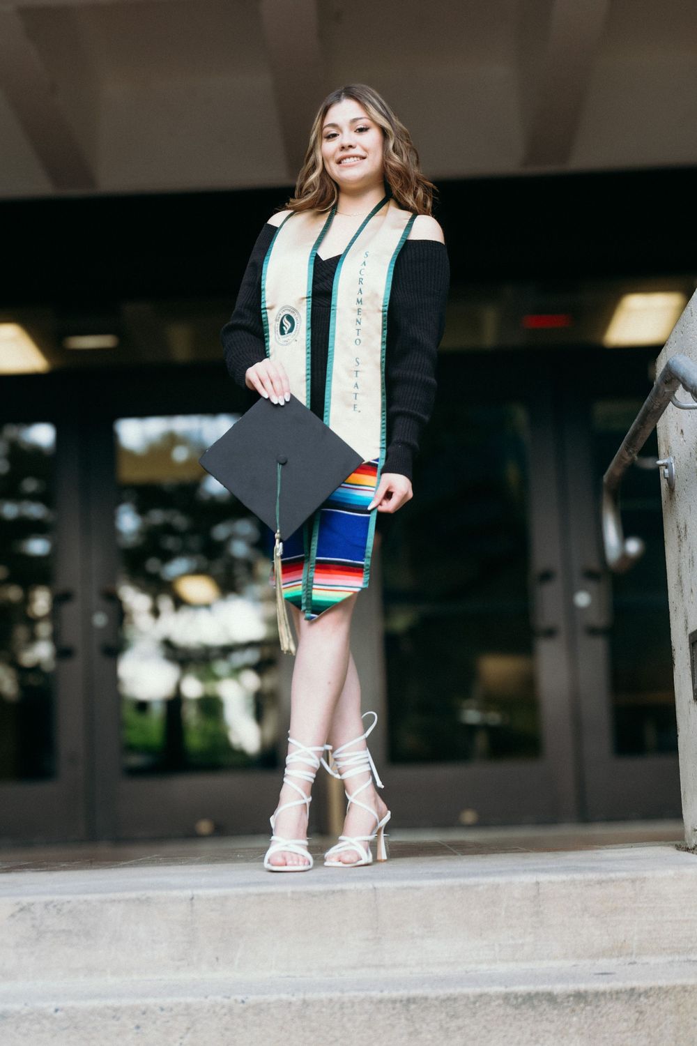 Young woman in colorful serape skirt holding graduation cap and wearing honor stole on university steps.
