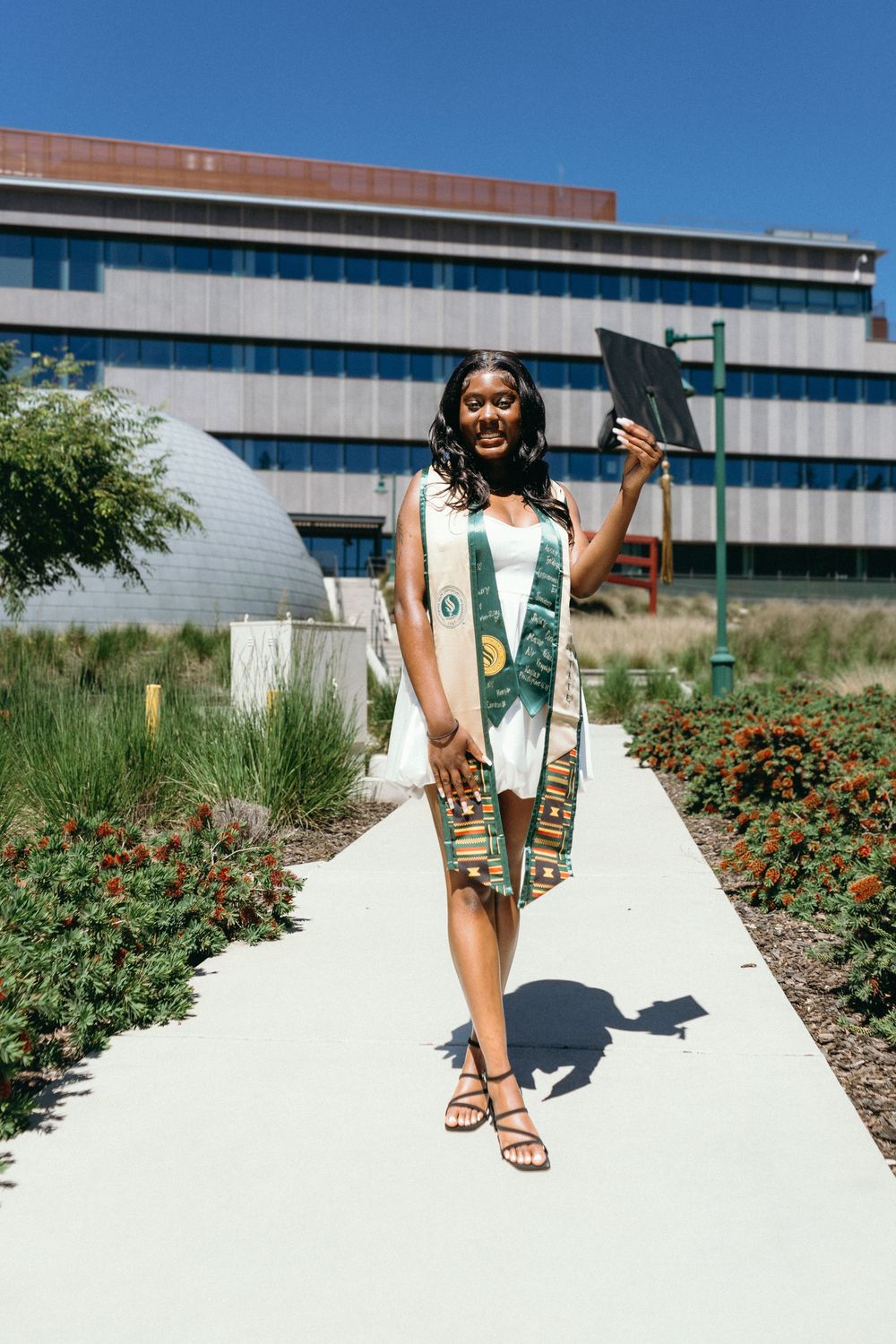 A smiling graduate in a white dress holds her cap while wearing a green stole on a sunny campus pathway.