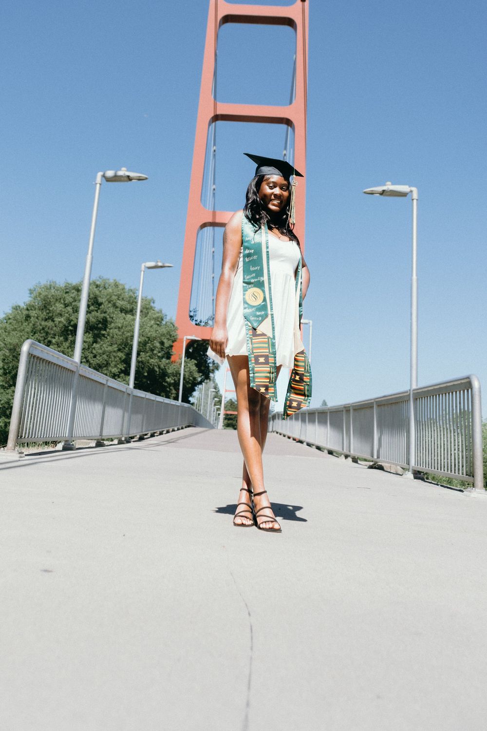 Young Black woman in graduation cap and sash poses proudly on the Golden Gate Bridge on a sunny day.