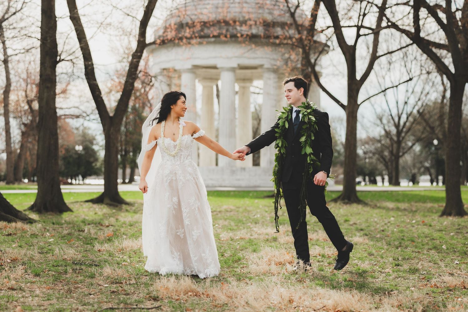 Bride and groom hold hands walking in a park with a classical rotunda monument in the background.