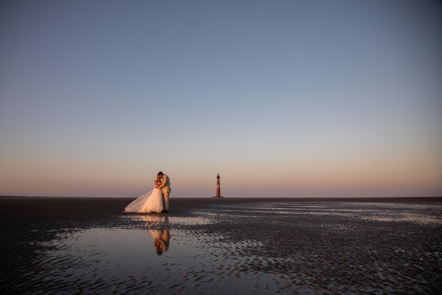 Bride and groom embrace on a reflective beach at sunset near a lighthouse.