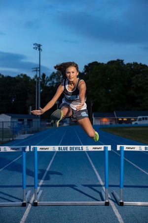Female high school athlete jumping over hurdles on a blue track.