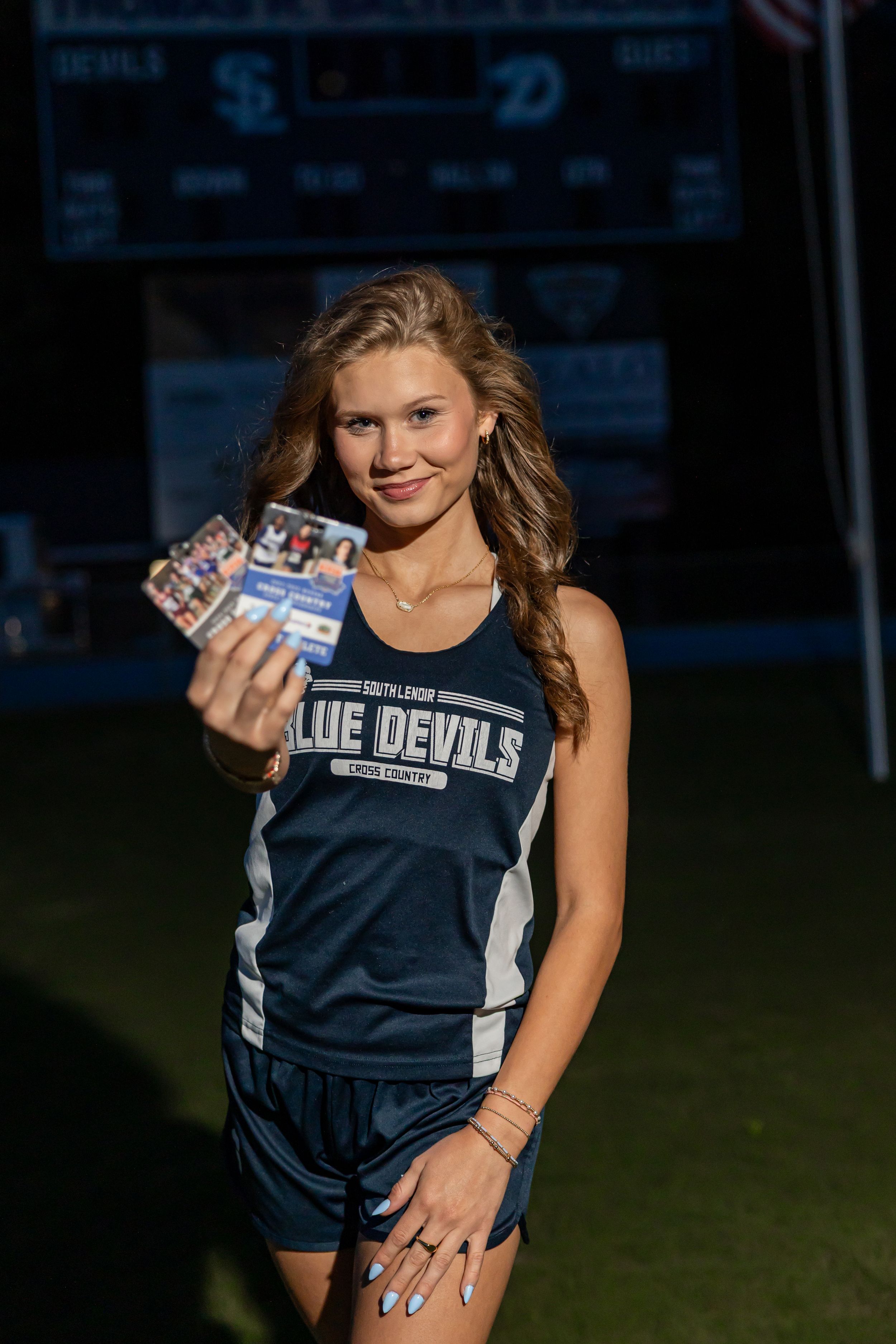 Young female athlete in Blue Devils cross country uniform smiling and holding trading cards on a sports field.