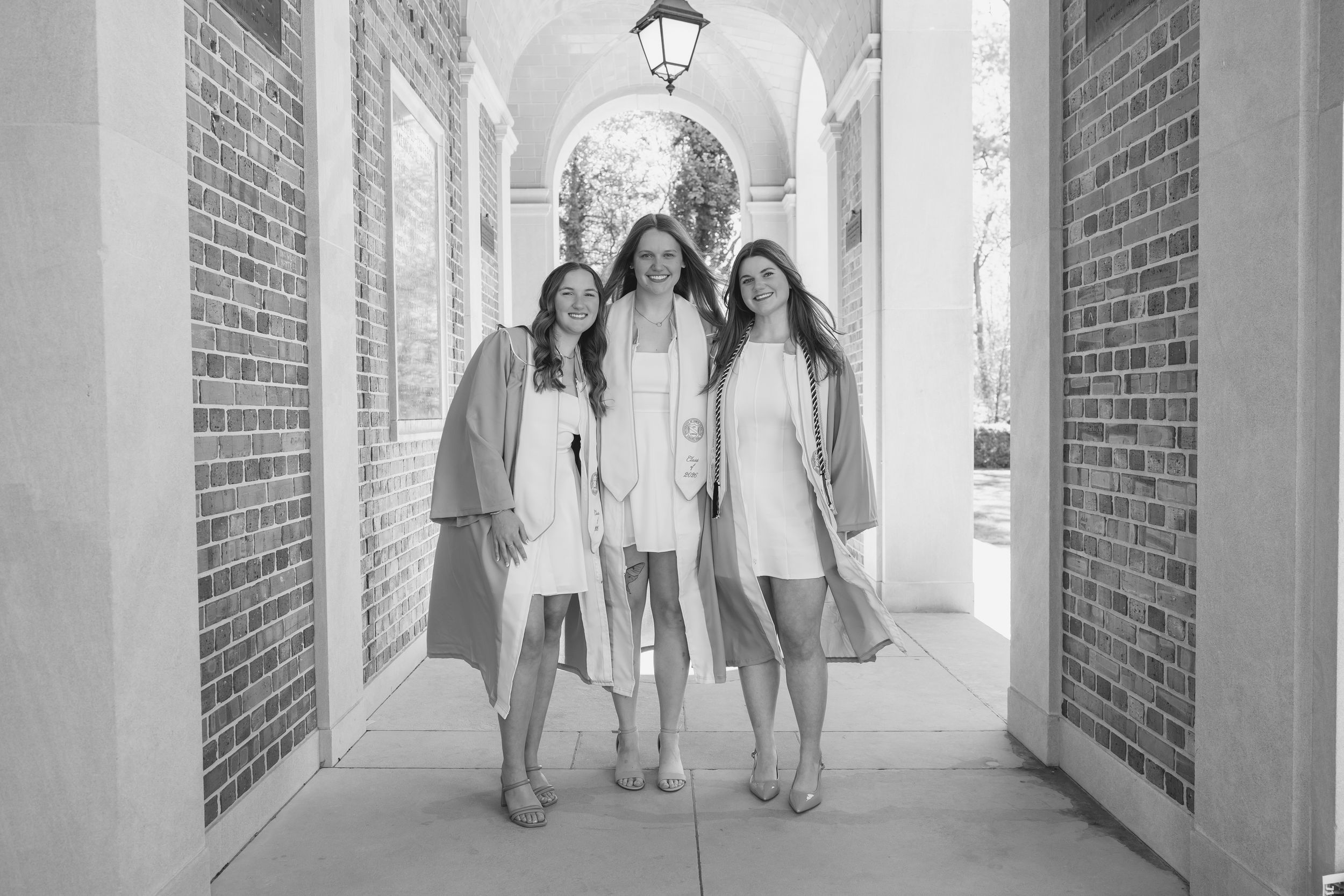 Three young women in white dresses and graduation robes smile together in a brick archway corridor.
