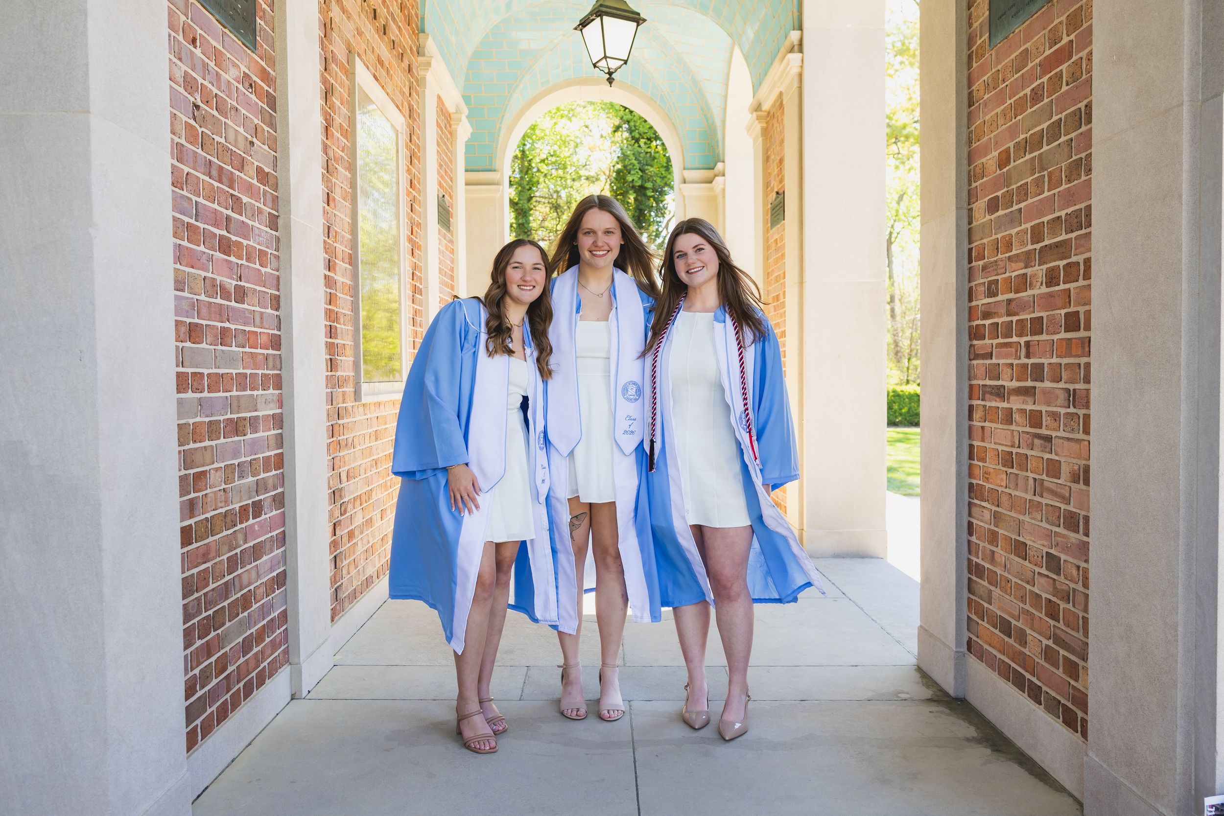 Three young women in blue graduation gowns smiling together in a brick archway corridor.