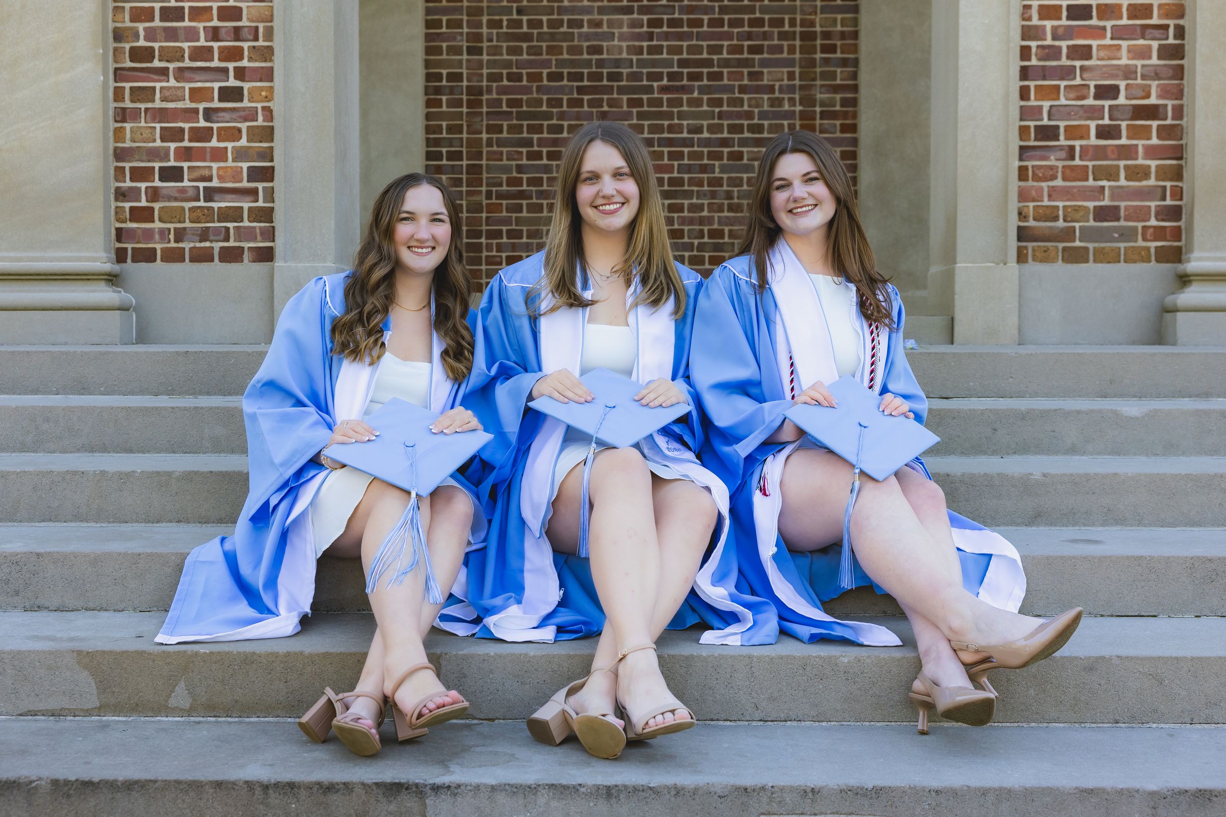 Three young women in blue graduation gowns sitting on stone steps, smiling and celebrating their academic achievement.