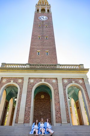 Three women in blue dresses sit on steps before a tall brick clock tower at a university campus.