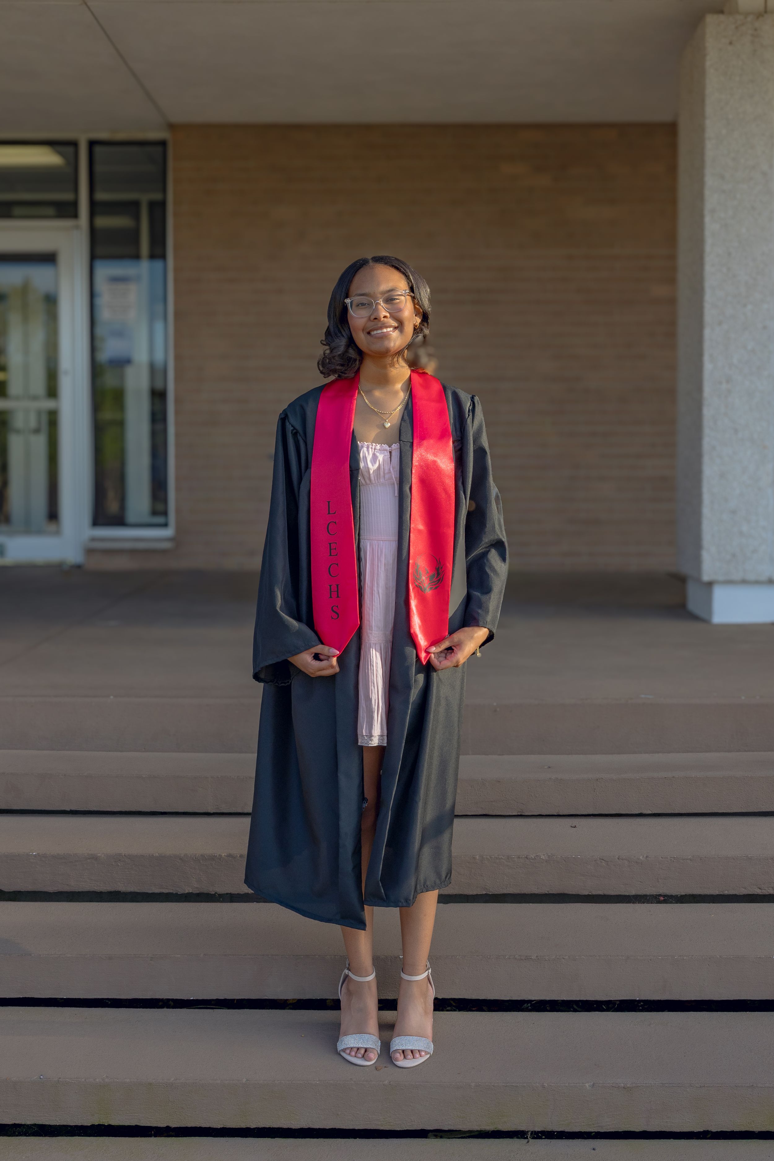 Young woman in black graduation gown with red sash smiling on steps outside a brick building.