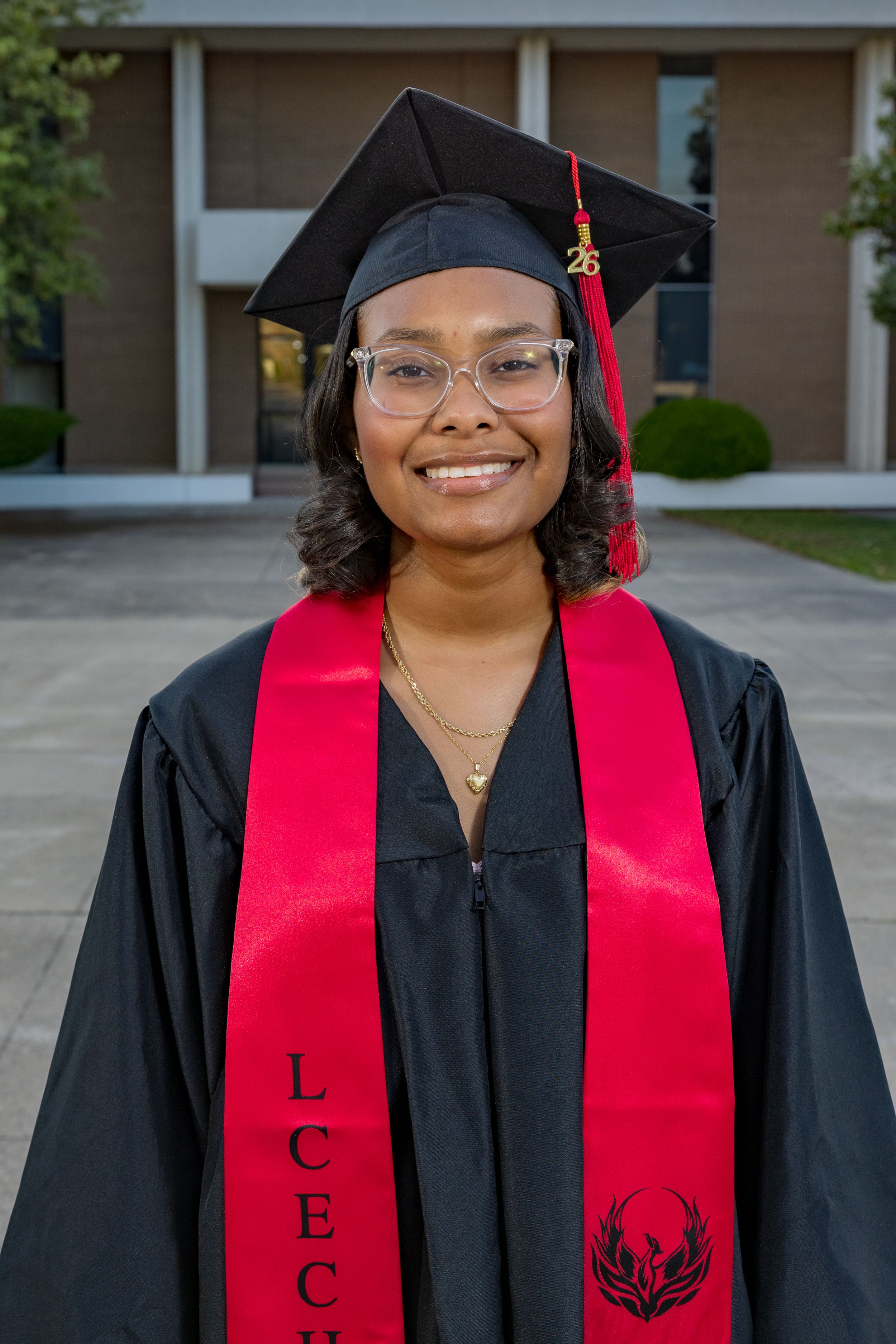 Smiling graduate in black cap and gown with red LCEC honor stole poses outdoors for graduation photo.