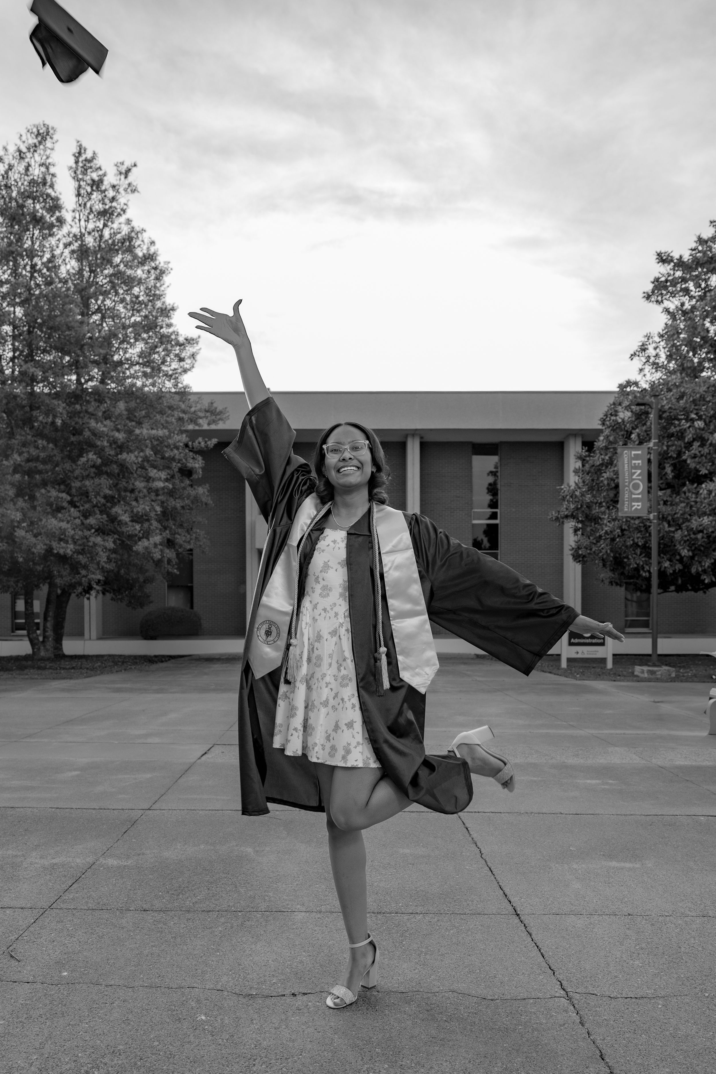 Black and white photo of a smiling graduate in cap and gown tossing her hat in the air outdoors.