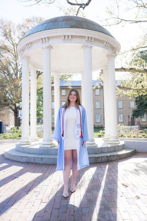 Young woman in light blue graduation gown stands before white rotunda columns on a sunny campus day.