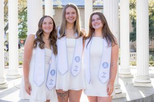 Three young women in white dresses and graduation stoles smile together in front of elegant white columns.
