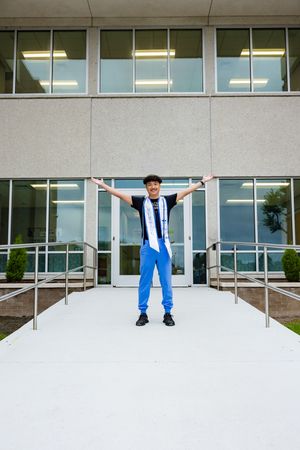 Young graduate in blue pants and stole stands arms wide open on campus ramp celebrating achievement.