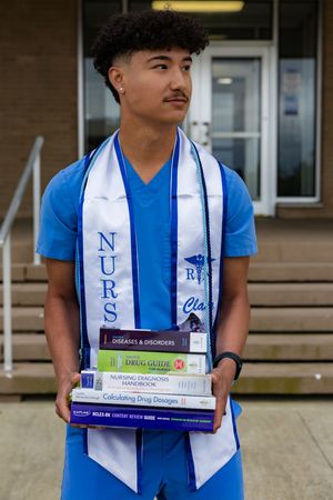 Young male nursing graduate in blue scrubs holds textbooks, wearing a NURS graduation stole outside a building.