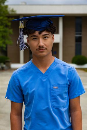 Young man in blue scrubs and graduation cap poses outdoors near a building, celebrating his healthcare degree.