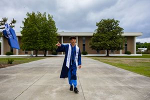 Graduate in blue cap and gown celebrates outdoors, waving regalia in front of a school building on a cloudy day.