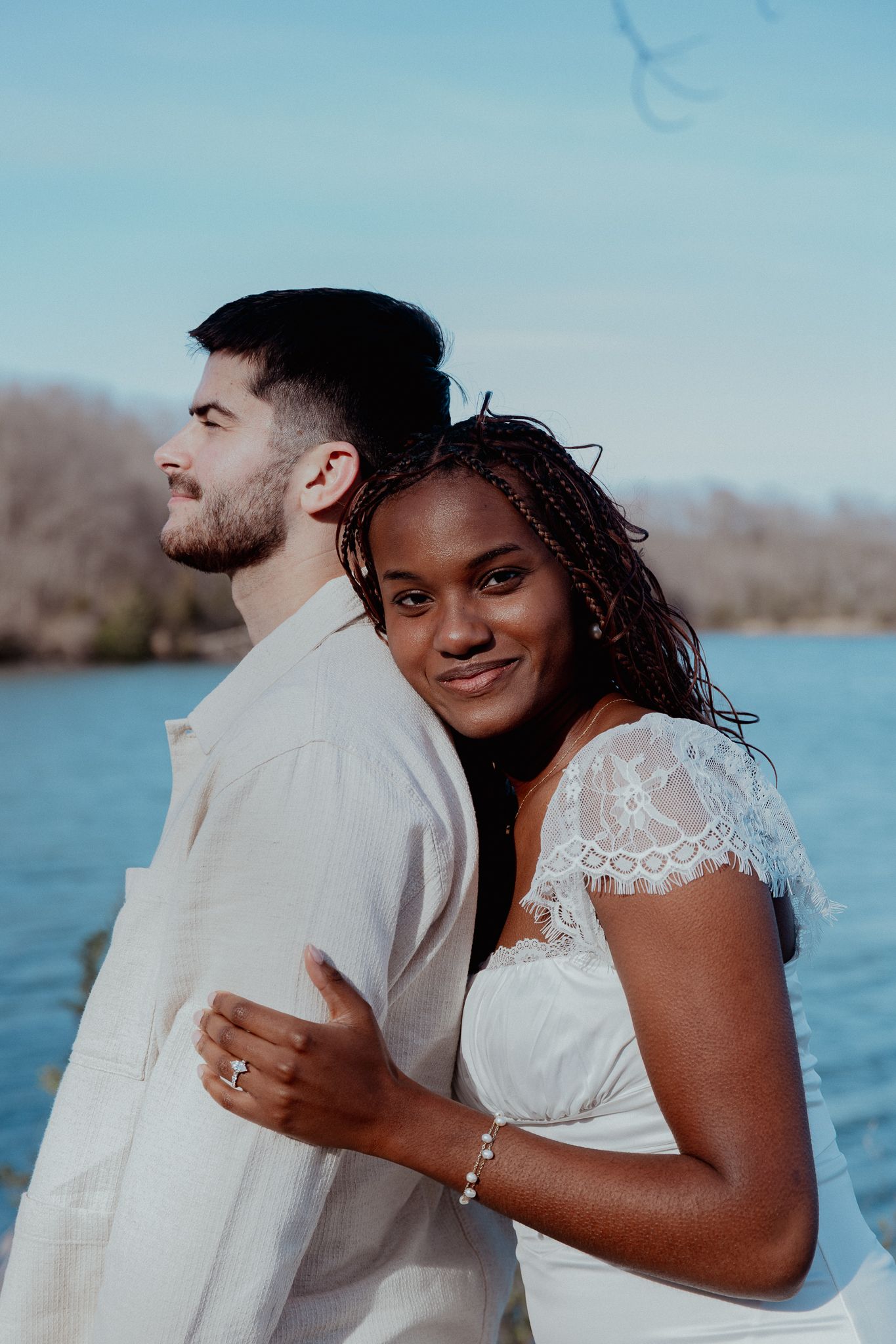 Interracial couple in white attire posing back-to-back near a lake, woman smiling at camera wearing engagement ring.
